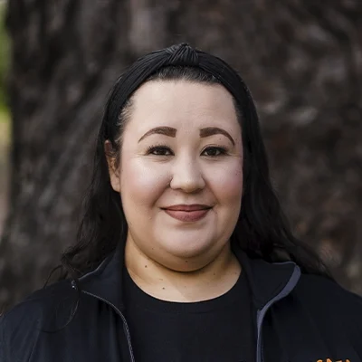 A headshot of Lucy wearing a black shirt, photographed outdoors with a tree background.