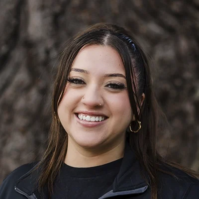 A headshot of Stephanie wearing a black shirt, photographed outdoors with a tree background.